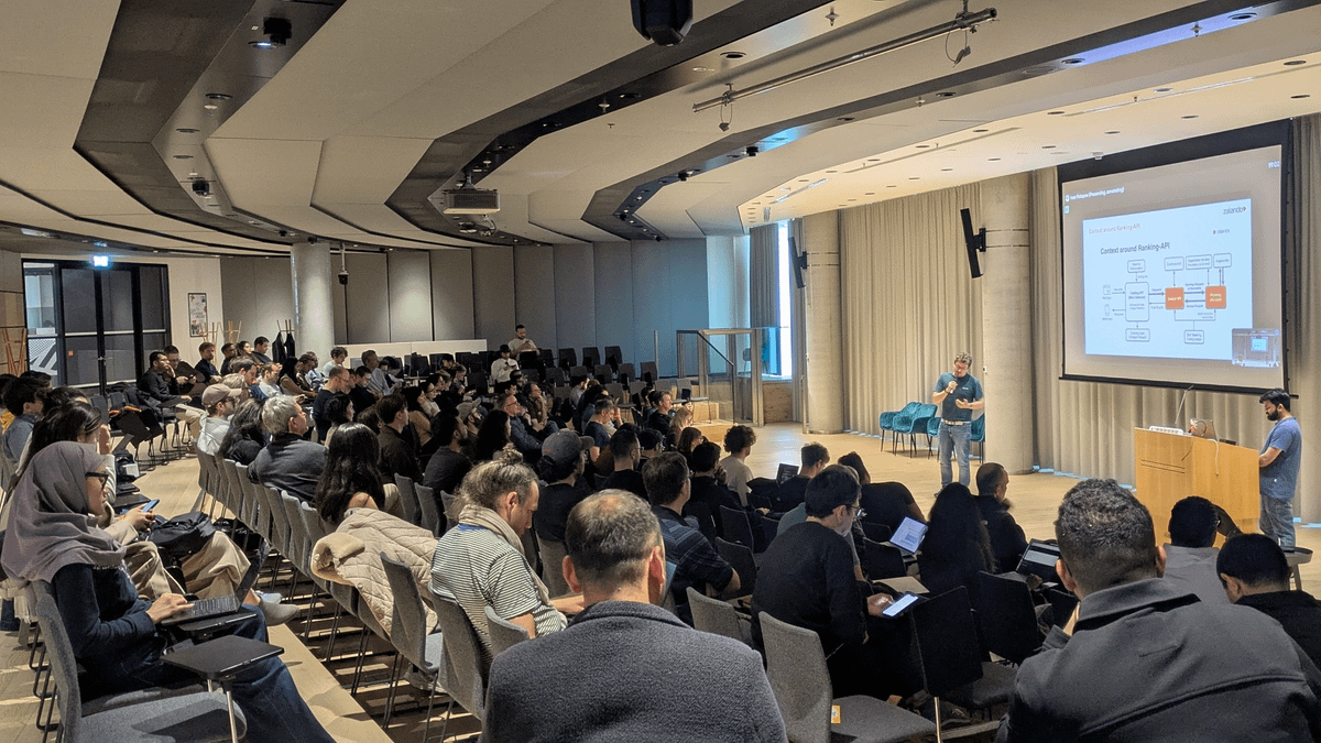 Attendees gather in a modern venue, listening to speakers discussing agentic search and workflows, with presentation visuals displayed behind them.