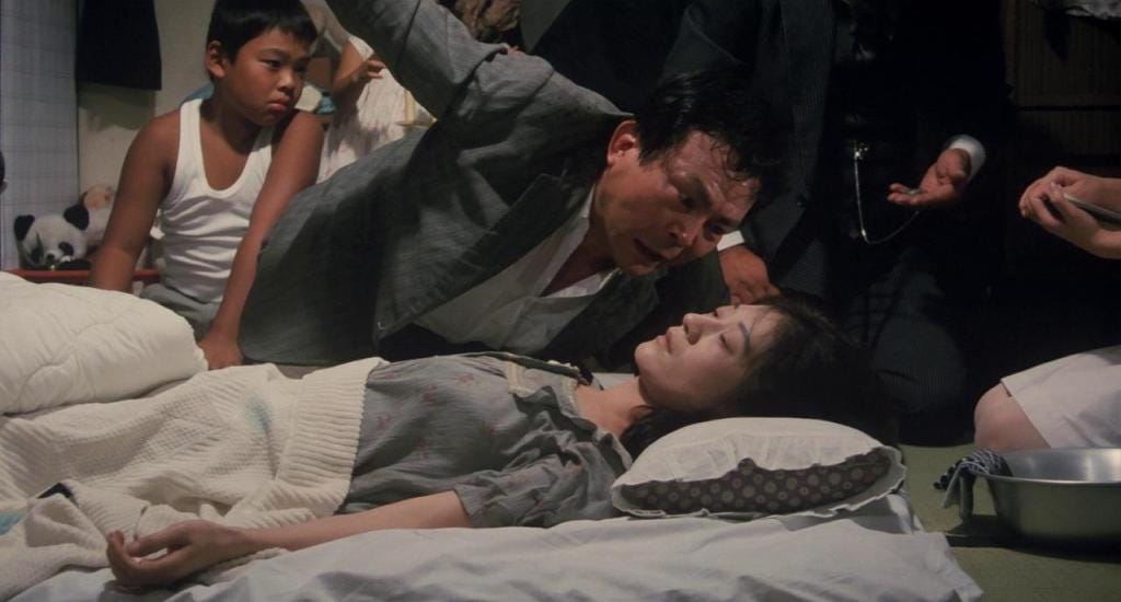 A tense scene shows a worried man leaning over a sickly woman lying on a bed, surrounded by concerned onlookers, conveying urgency and distress.
