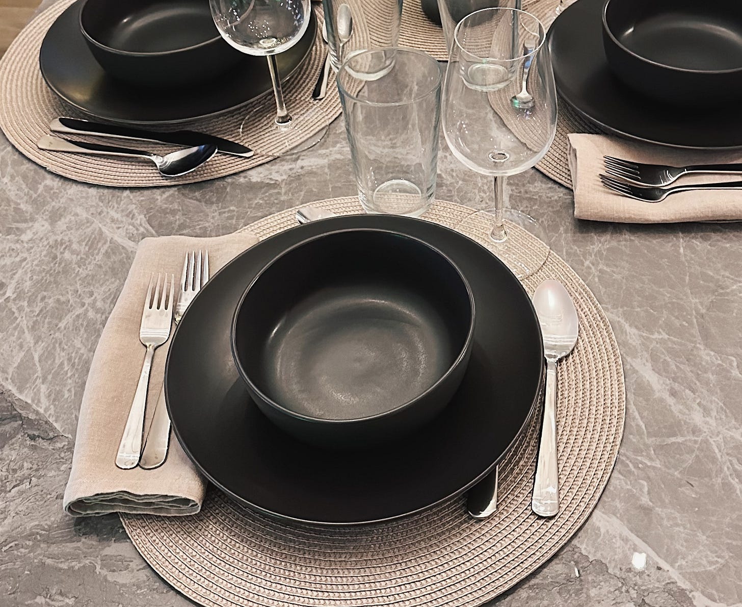 Neutral linen napkins on a gray stone dining table with woven placemats, matte gray ceramic dinnerware, clear glassware, and silver flatware.