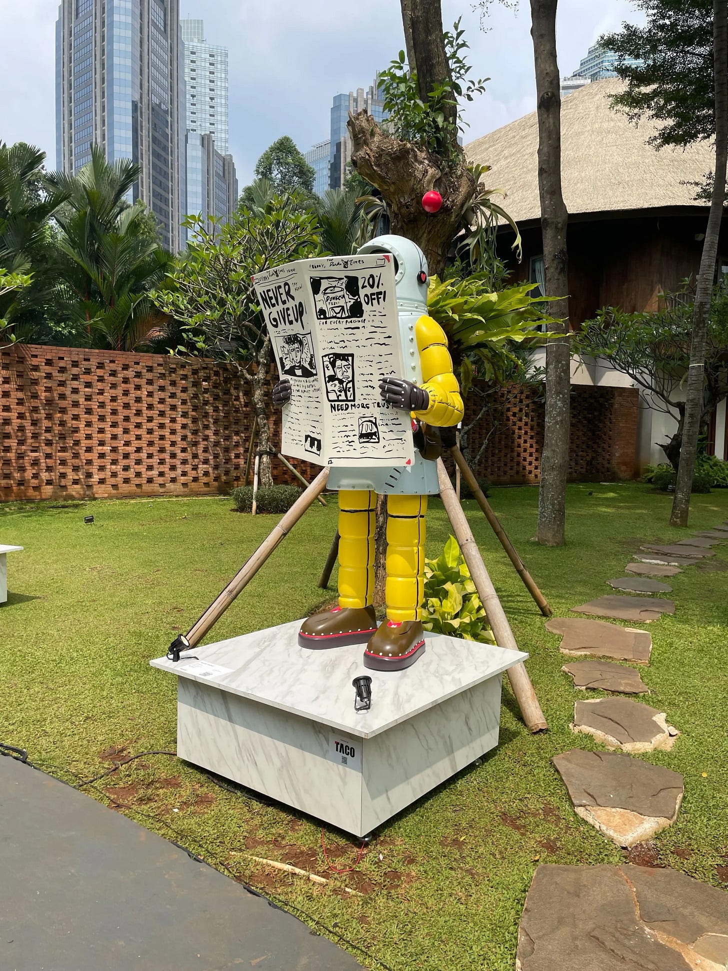 A large colourful robot sculpture standing on a marble plinth in a lush tropical garden, holding an oversized newspaper with the text "Never Give Up" visible on the front page. The robot has a light blue torso, yellow articulated arms and legs, brown boots, and a red antenna on its head. Jakarta skyscrapers and palm trees visible in the background. Part of Art Jakarta.