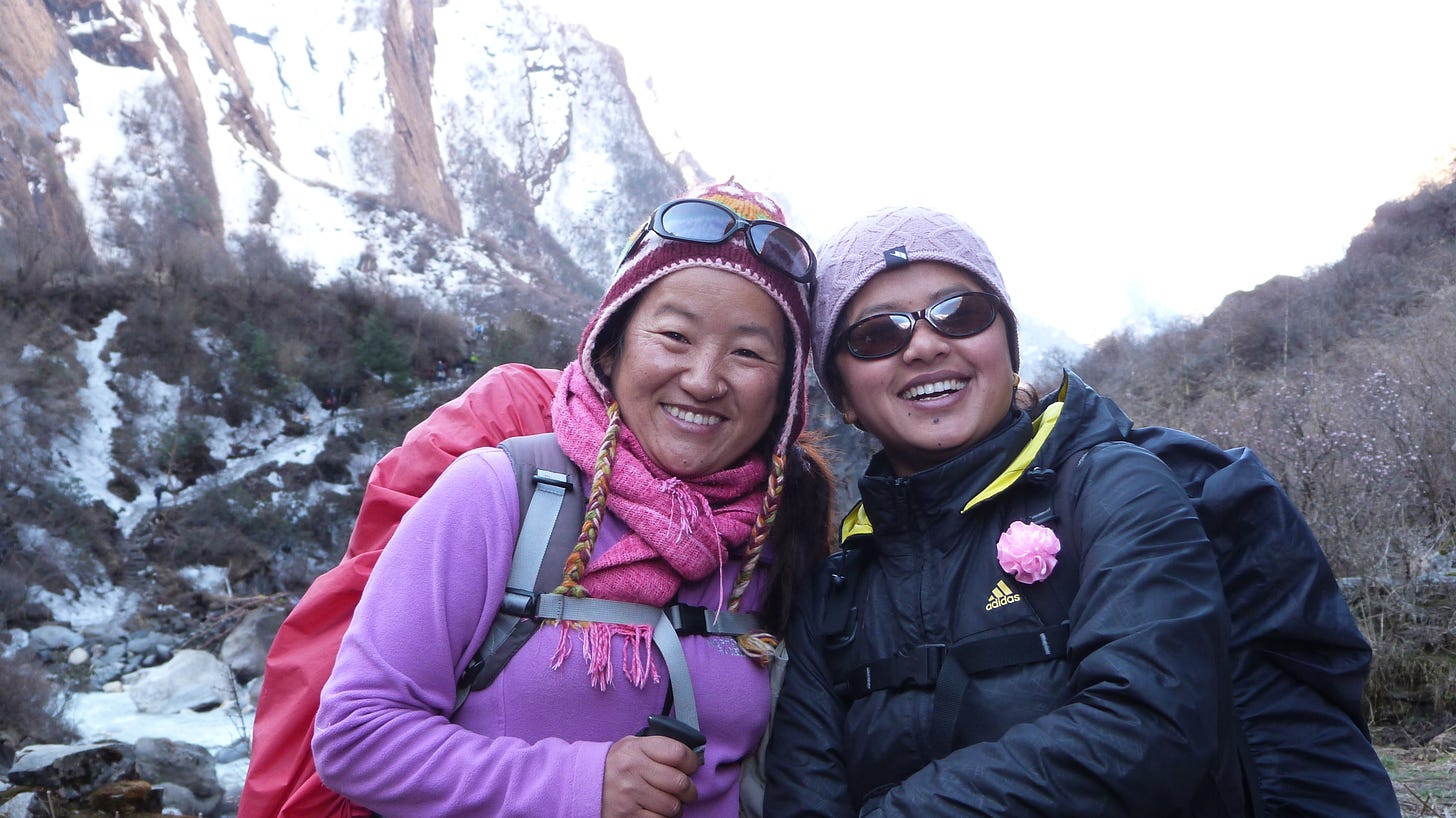 Two Nepali women are wearing cold-weather gear. One is in pink and the other dark blue. They are wearing backpacks and smiling while hiking in the mountains.