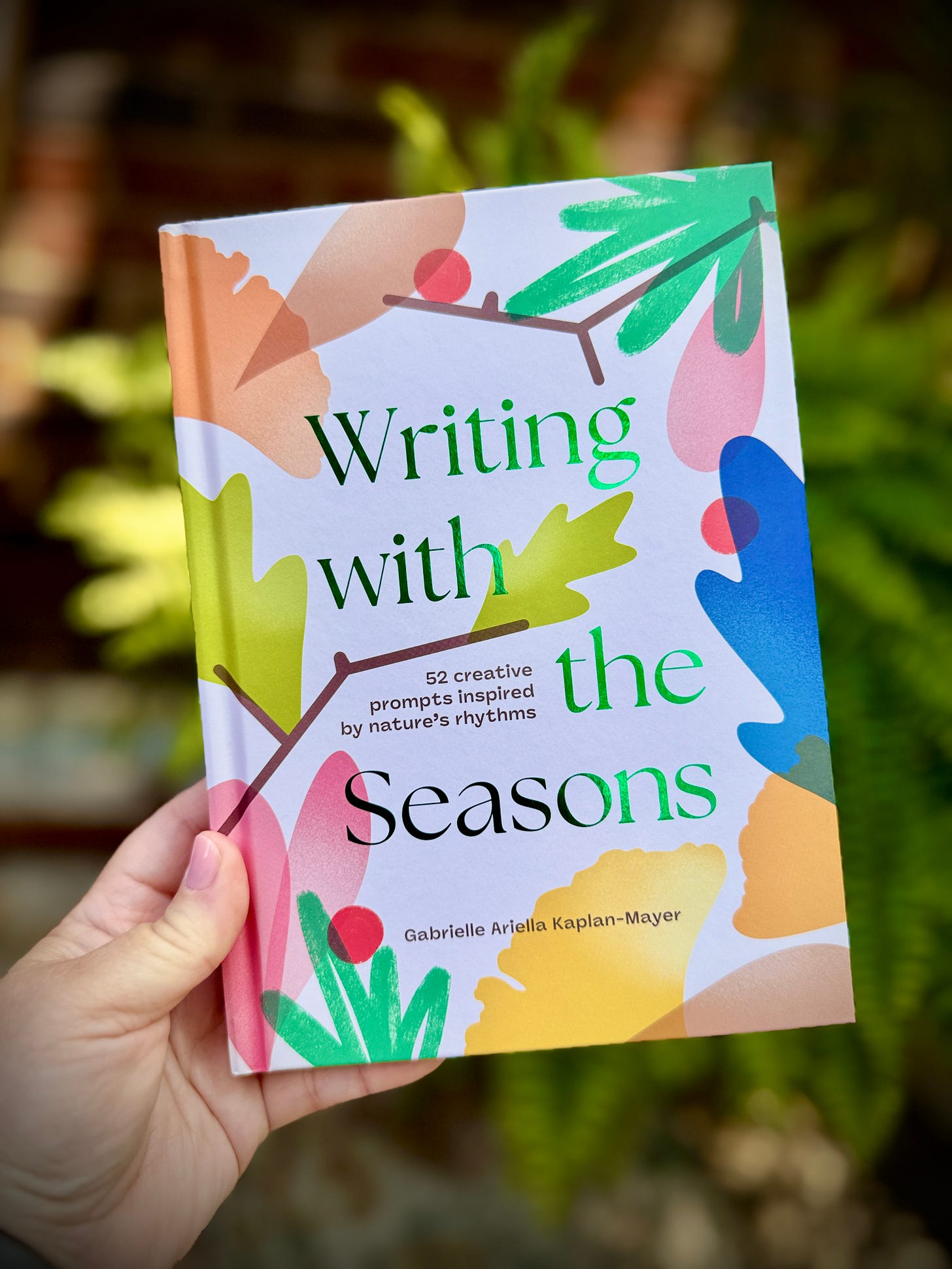 A hand holding up the cover of the journal Writing With The Seasons: 52 creative prompts inspired by nature's rhythms by Gabrielle Ariella Kaplan-Mayer against a soft green leafy background.