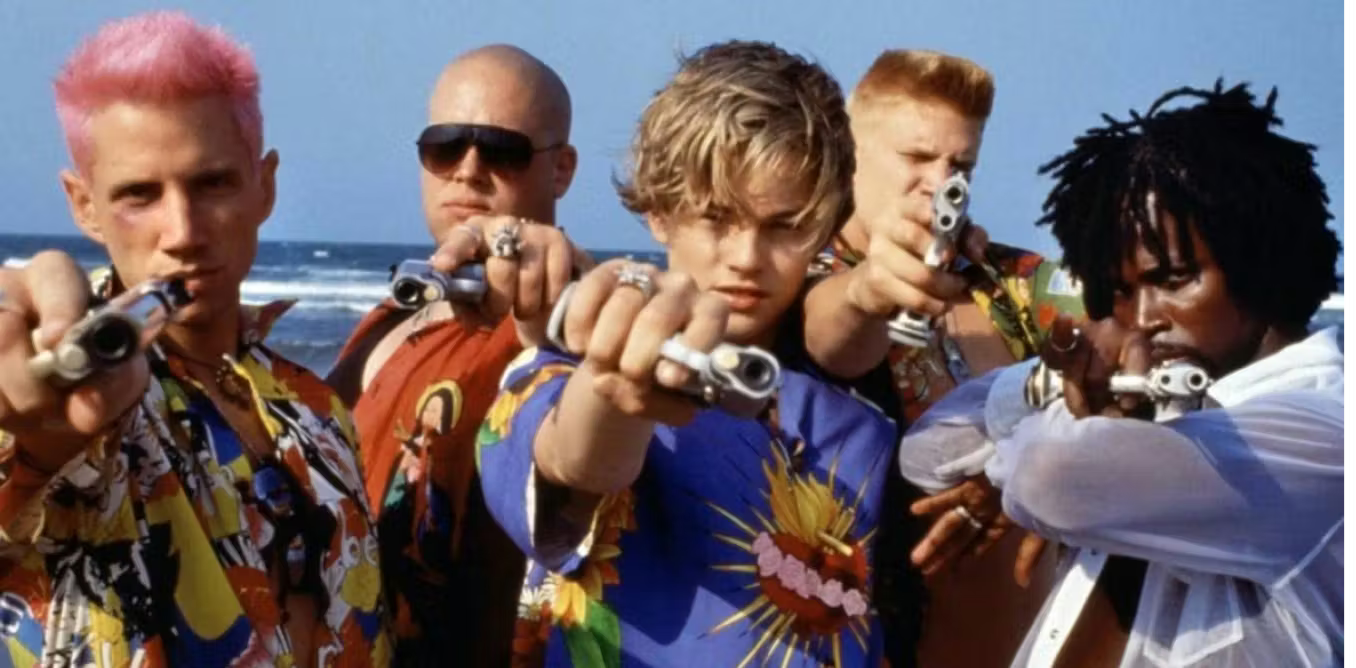 Still from Baz Luhrmann's Romeo & Juliet: five men on a beach in colorful religious-print shirts pointing guns at the camera.