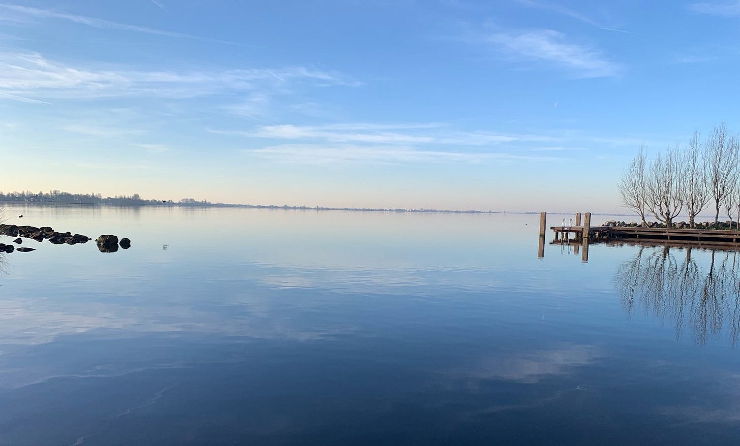 Calm lake with a wooden pier on the right, reflecting the clear blue sky and distant trees.