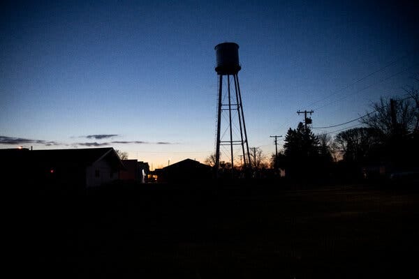 A water tower in shadow at sunset over a horizon of small buildings, trees and power lines.