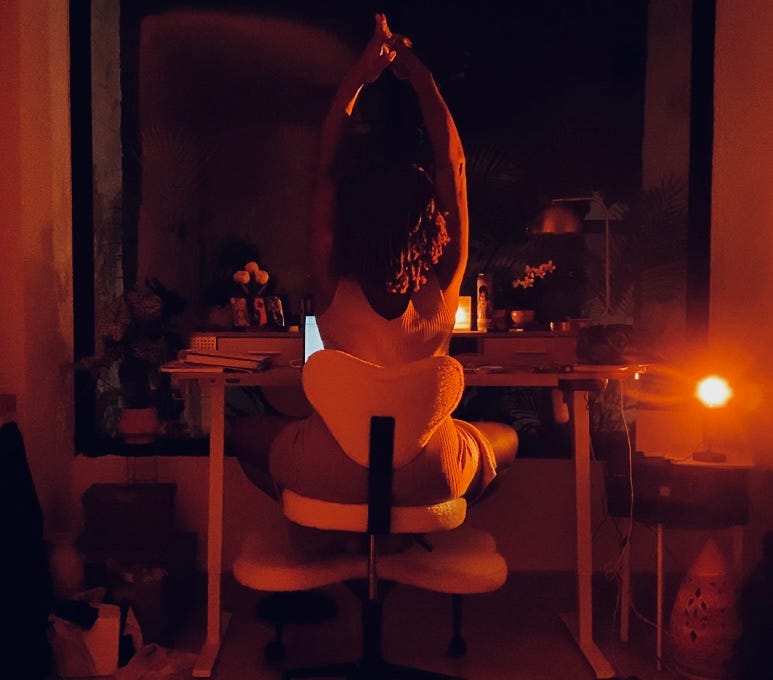 A Black woman sitting in a sculpted white desk chair at night, arms stretched overhead, lit by candlelight and a small orange lamp. Her laptop glows on the desk in front of her, surrounded by plants and altar objects, creating a sensual, focused workspace. A Black woman sitting in a sculpted white desk chair at night, arms stretched overhead, lit by candlelight and a small orange lamp. Her laptop glows on the desk in front of her, surrounded by plants and altar objects, creating a sensual, focused workspace.