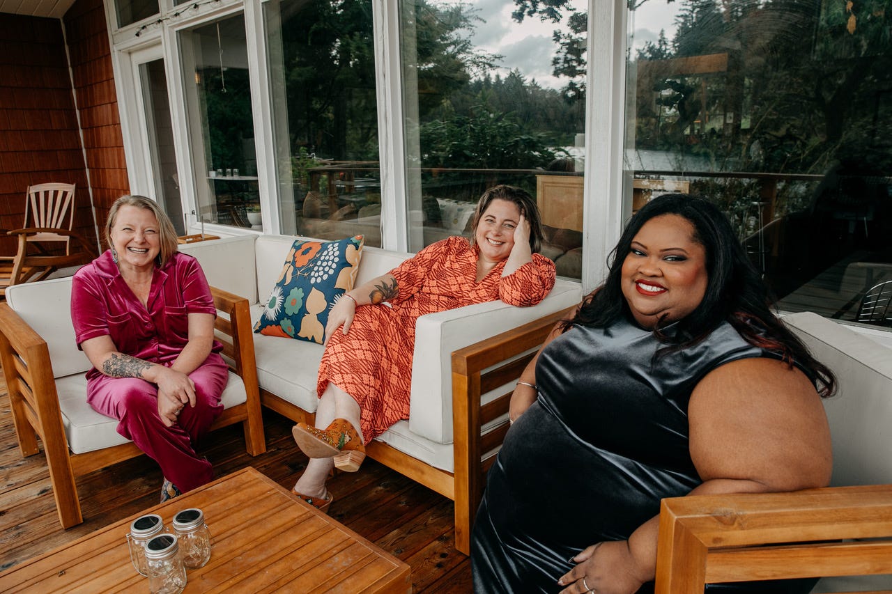 Three women sit on a wooden deck on wood framed furniture with white cushions and a wall of windows behind them. Dana Sturtevant, a white woman with chin length blonde hair, wears a bright pink velour pants and shirt, Hilary, a while woman with shoulder length brown hair wears an orange dress with a plaid print, Sirius, a Black woman with long black hair wears a black satin dress, all are smiling. Three women sit on a wooden deck on wood framed furniture with white cushions and a wall of windows behind them. Dana Sturtevant, a white woman with chin length blonde hair, wears a bright pink velour pants and shirt, Hilary, a while woman with shoulder length brown hair wears an orange dress with a plaid print, Sirius, a Black woman with long black hair wears a black satin dress, all are smiling.