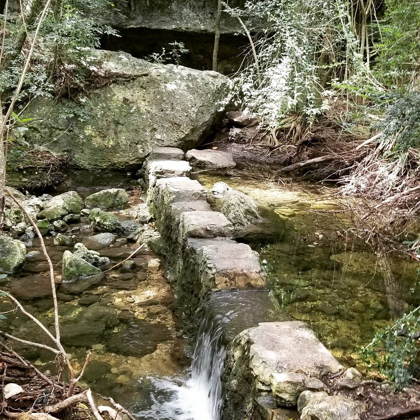 A clear creek with a short dam. Water is flowing over the dam to another poll about eon foot below. Across the dam are crenelations serving as slepping stones. IN the background are large stones with a cave-like entrance.