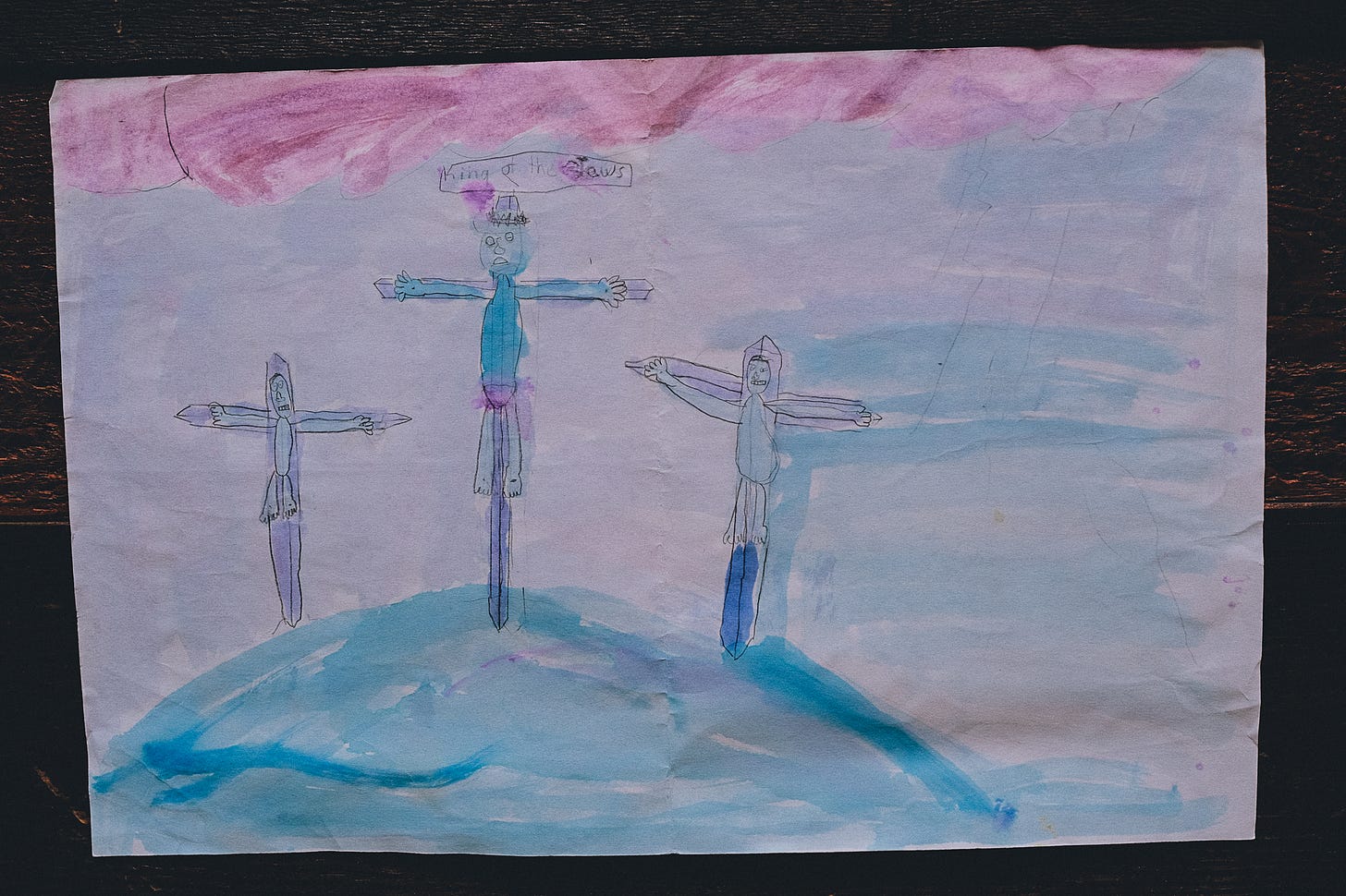 a watercolor painting by a child of three people on crosses looking very unhappy. above the one in the middle is a sign that says king of the jews