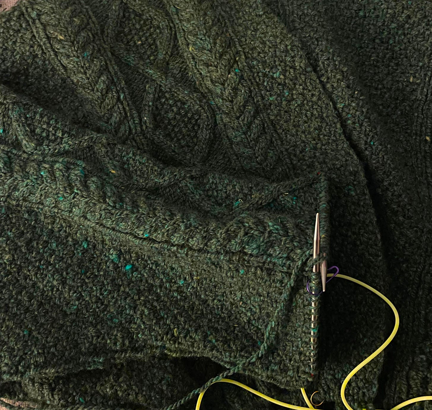 An image of a dark green heavily cabled cardigan. There are bright yellow cords and an in-progress sleeve featured.