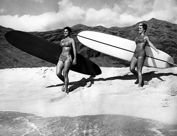 Two women carrying their surfboards along the beach, 1960. Two women carrying their surfboards along the beach, 1960.