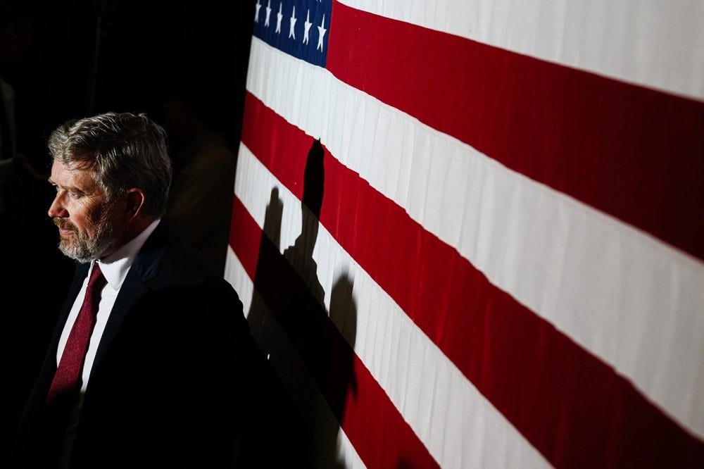 Thomas Massie, seen at profile, with dramatic lighting, which casts a hard shadow on the large American flag behind him (on the right side of the frame).
