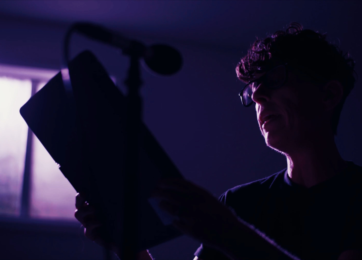 Andrea Gibson in a purple dimly lit room reading in front of a microphone and holding a book. Andrea is wearing glasses. The tone is somber.