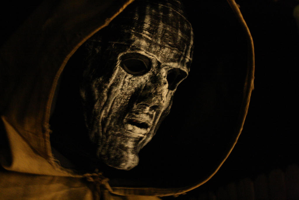 uma imagem de alguém usando uma máscara de material desconhecido, de cor branca com vincos, um capuz amarelo, ao redor da imagem há apenas a cor preta, assim como nos olhos e por dentro do capuz.