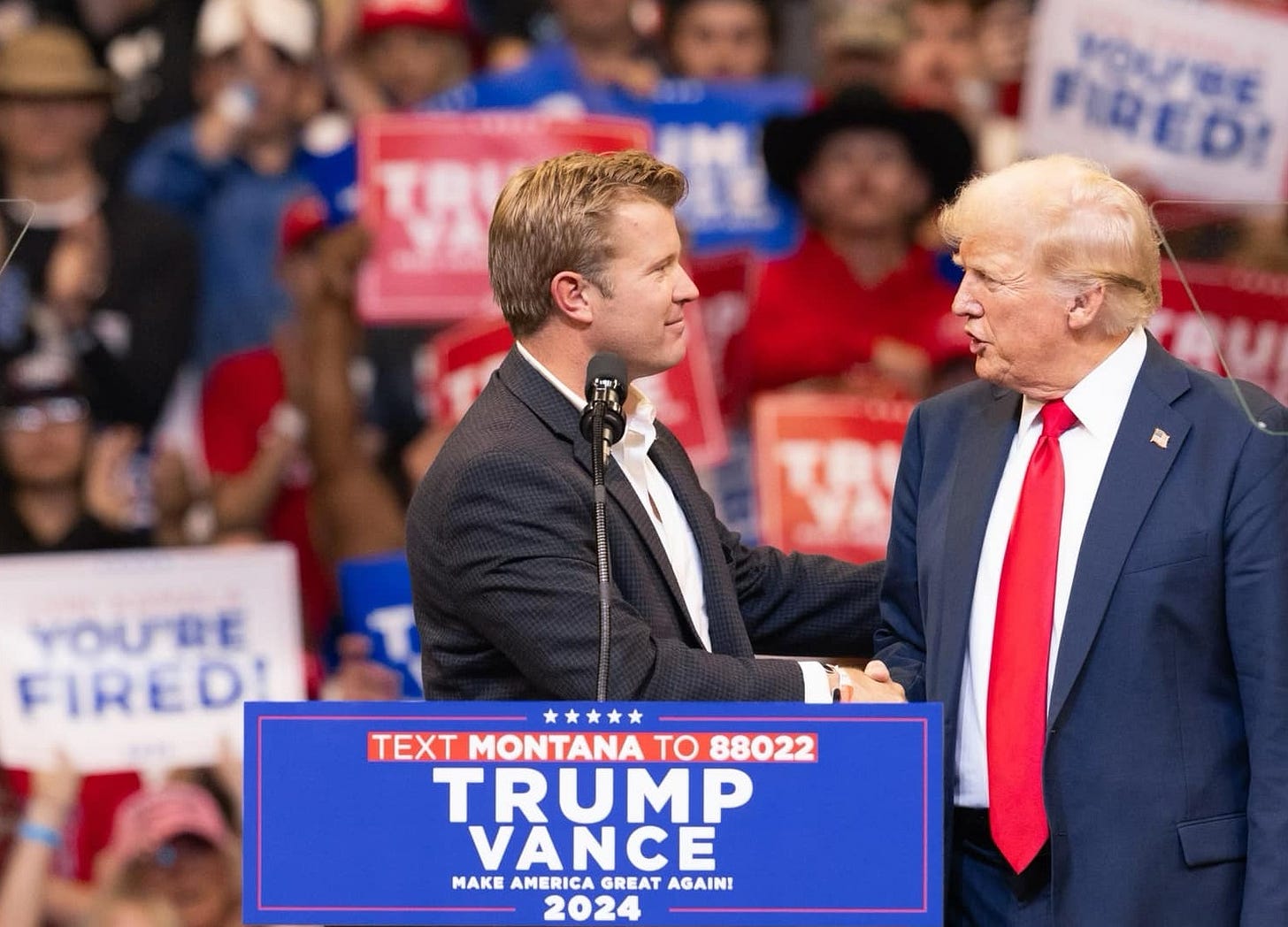 Senate candidate Tim Sheehy shakes Donald Trump's hand at a rally. In the background, two members of the audience wave signs reading 'YOU'RE FIRED' Senate candidate Tim Sheehy shakes Donald Trump's hand at a rally. In the background, two members of the audience wave signs reading 'YOU'RE FIRED'