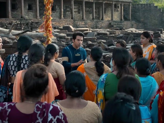 A still from Hindi film, ‘Stree 2’. Vicky (Rajkummar Rao), wearing a dark blue-green hoodie, speaking to a large-ish group of women. A ruin of sorts with pillars makes the background.