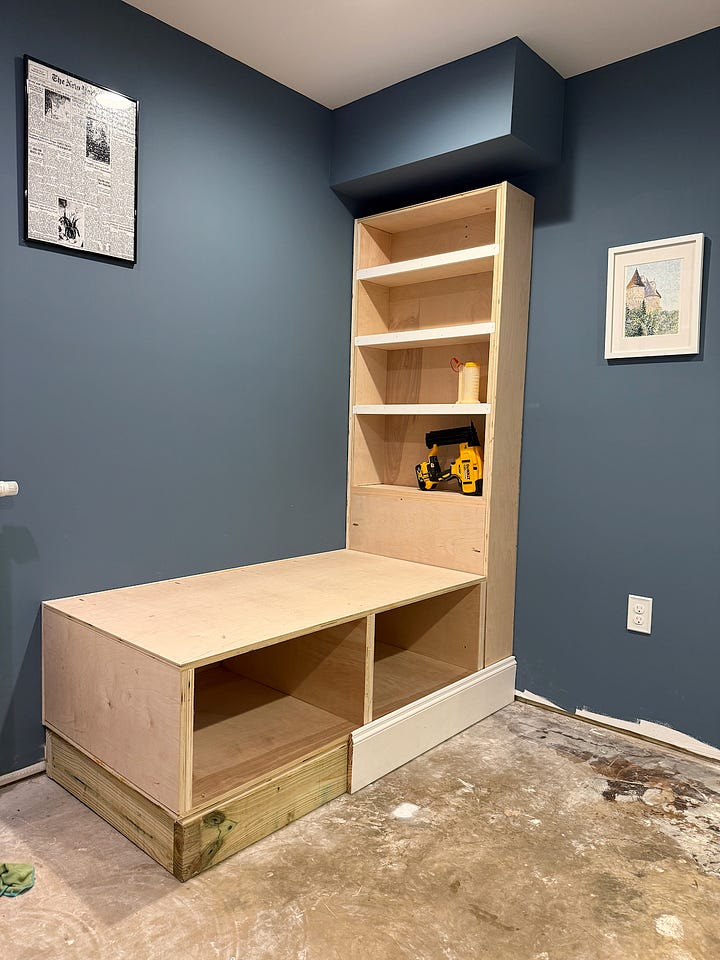 Alt text: A four-photo collage documenting the construction of a custom built-in reading nook. The top-left image shows an empty corner of a room with blue walls, white trim, and a ceiling bulkhead. The top-right image shows the initial framework for a bench seat and a vertical cabinet side installed beneath the bulkhead. The bottom-left image shows the bench structure further along, with additional framing and support panels in place. The bottom-right image shows the nearly completed project, including a built-in bench with storage compartments below and a floor-to-ceiling bookshelf fitted beneath the bulkhead. Together, the images illustrate the progression from an empty corner to a custom built-in seating and storage feature.