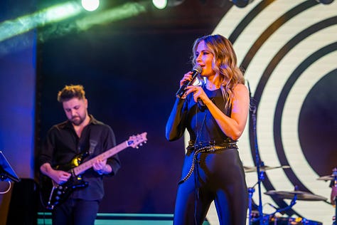Alina Crișan & Keo doi artiști cântă împreună pe scenă, față în față, fiecare ținând câte un microfon. Artista poartă o ținută neagră, iar artistul poartă o geacă din piele. Fundalul scenei are elemente grafice luminoase în tonuri de verde și galben, iar atmosfera este specifică unui concert live.