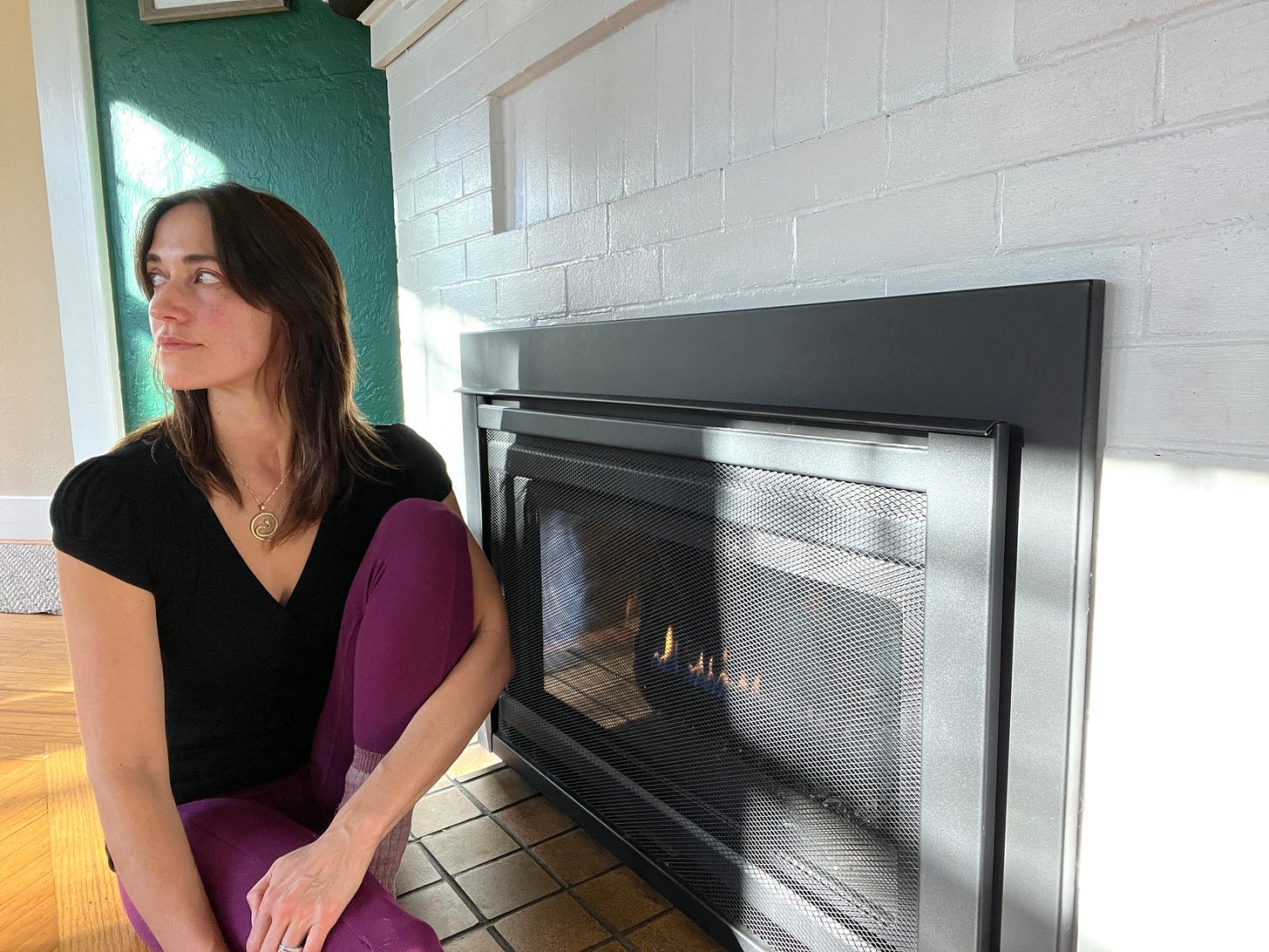 Rayann sits on the floor beside the fireplace, inches away from it. Her face is a little flushes from the warmth. She is looking out the window and morning light comes in.