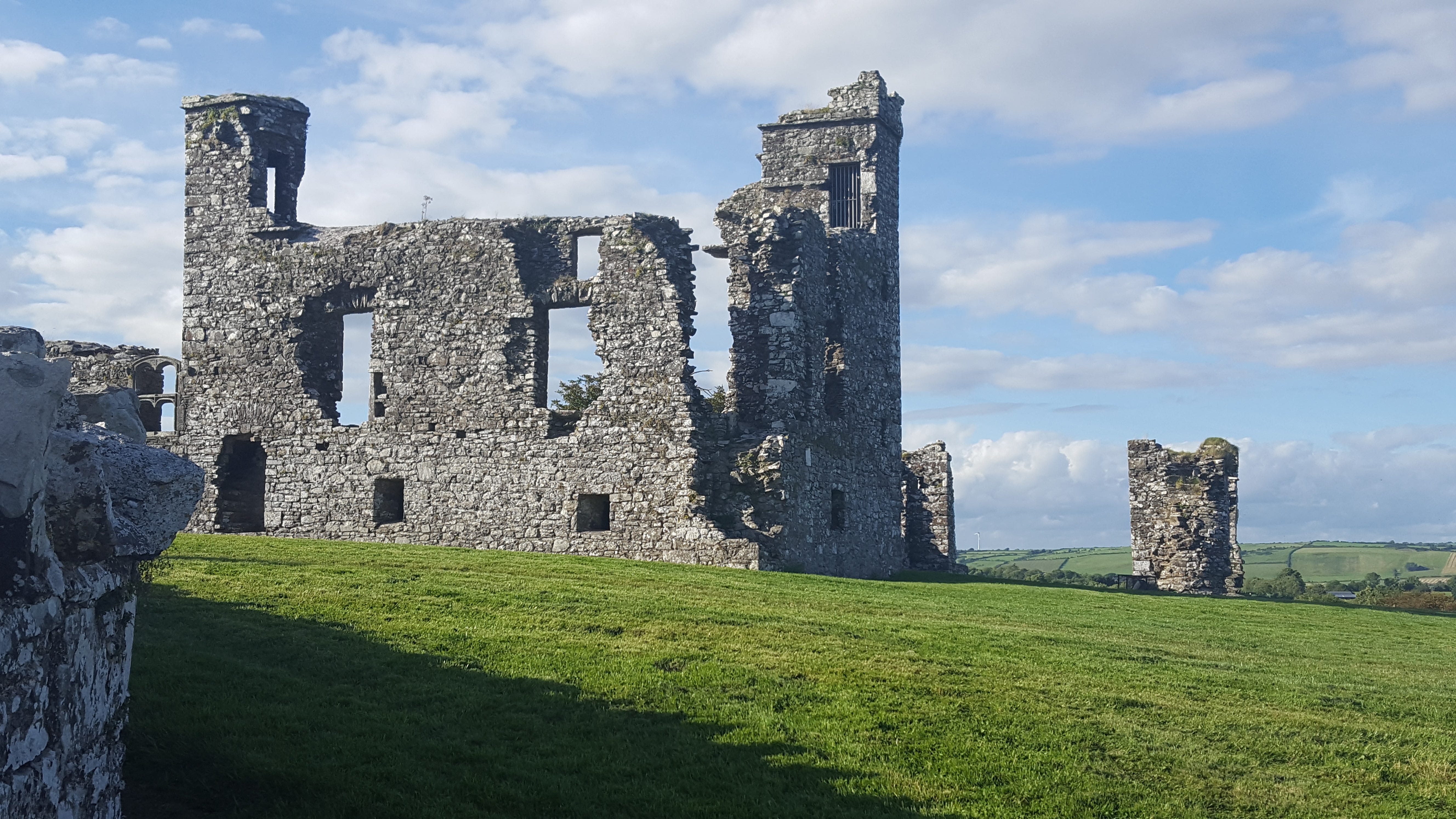 Grey ruined stone building of an abbey, well-weathered and tumbled, against a blue sy with drifting clouds on a sunny day.