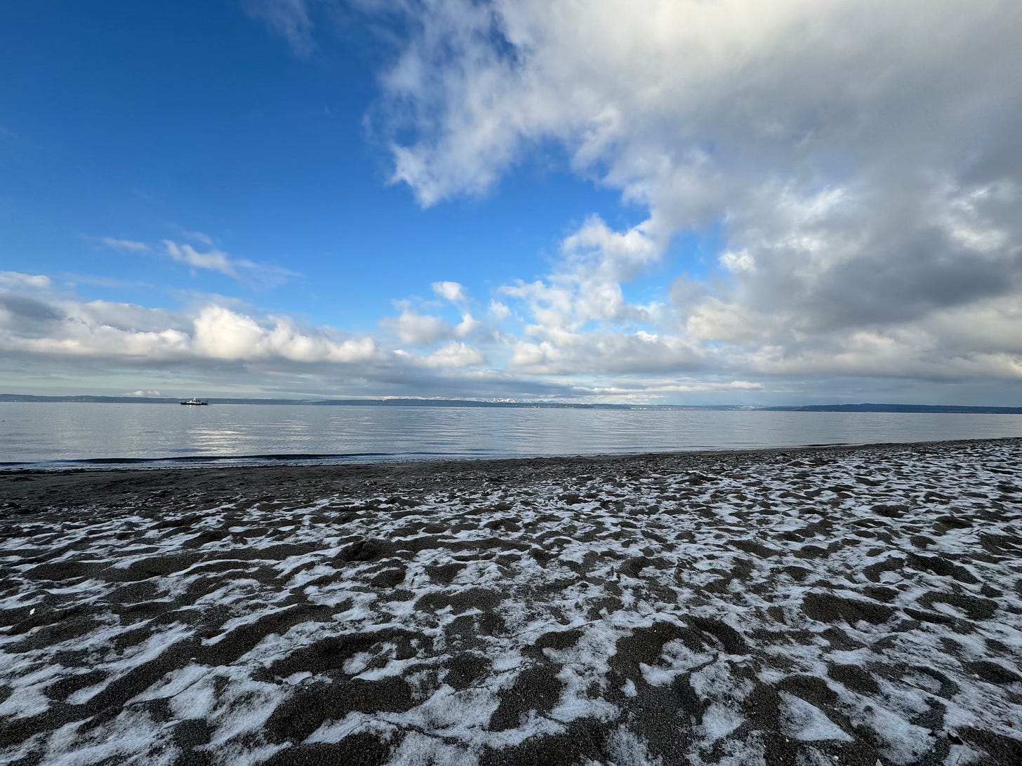 A wide beach view at Golden Gardens Park in Seattle showing dark sand covered with patches of white snow in intricate patterns. The calm Puget Sound stretches to the horizon under a cloudy Pacific Northwest sky, with distant mountains barely visible across the water.
