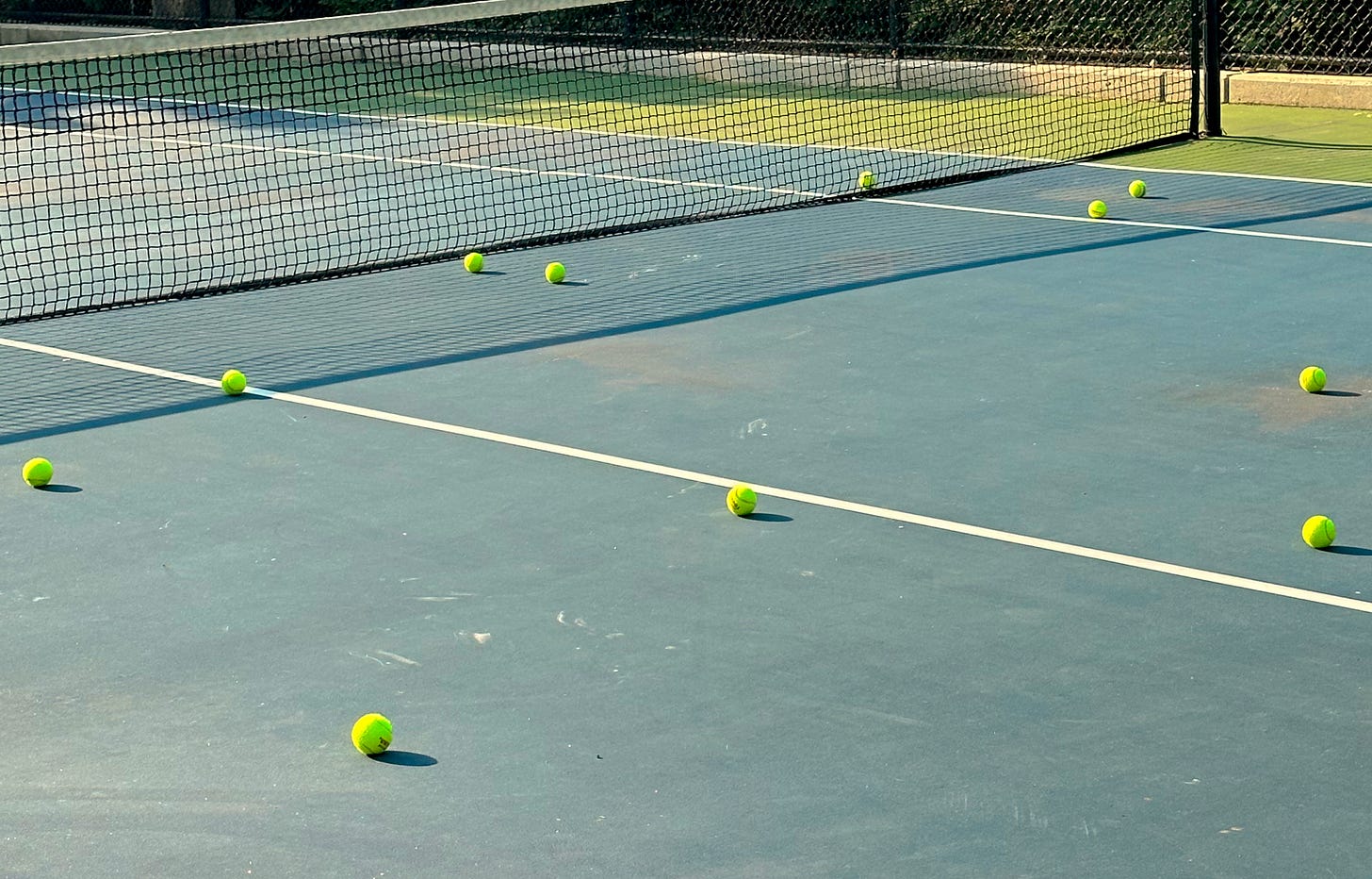 A tennis court with multiple bright yellow tennis balls scattered near the net, indicating a practice session. The court has a blue surface with white boundary lines, and a black net stretches across the middle.