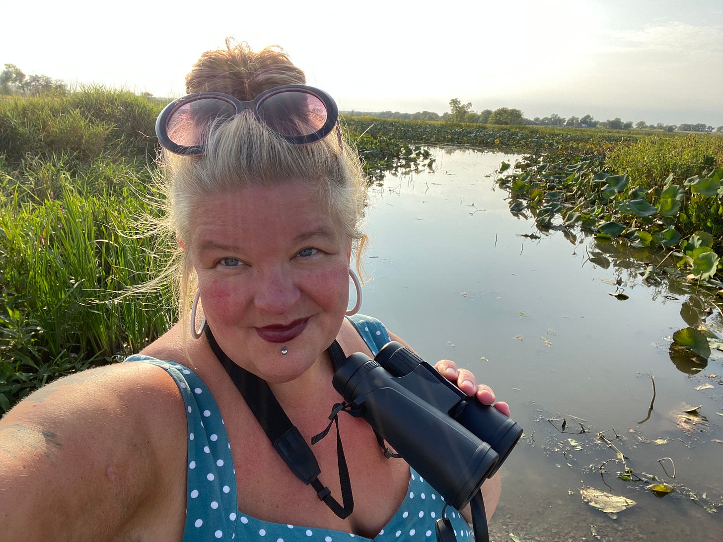 Cat takes a selfie in the early morning light with a placid meandering slough in the background. The banks are lined with large glossy green lily pads and other vegetation. Cat sports a blue top with white polka dots, dark red lipstick, and a silver stud beneath her lips. Her large round purple-tinged glasses rest on her blonde hair while she grasps a pair of binoculars with one hand.