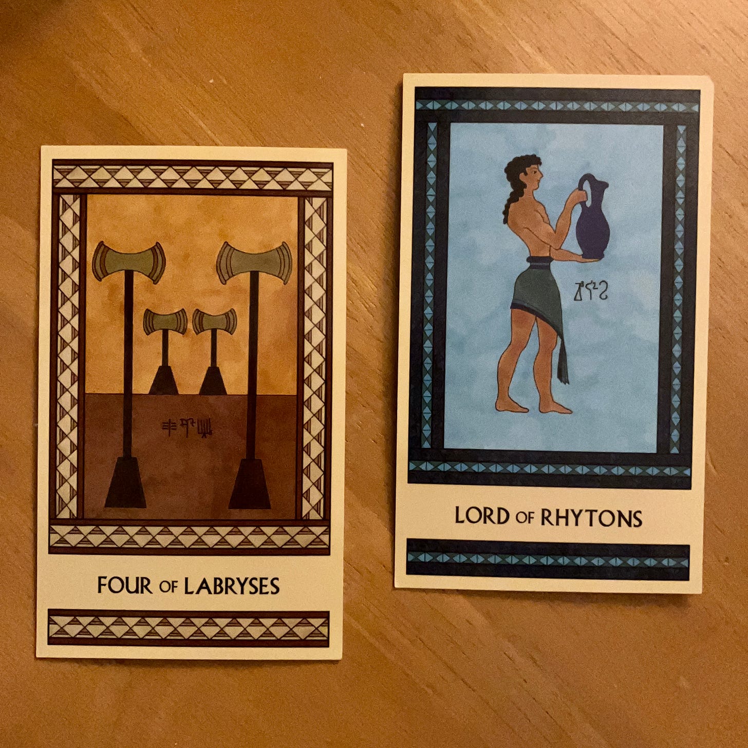Two Minoan Tarot cards side by side on a golden wood surface. The Four of Labryses shows two short gold labryses and two tall ones standing peacefully in a quiet place. The Lord of Rhytons shows a tan-skinned Minoan man wearing a blue-green kilt and carrying a large blue pitcher.
