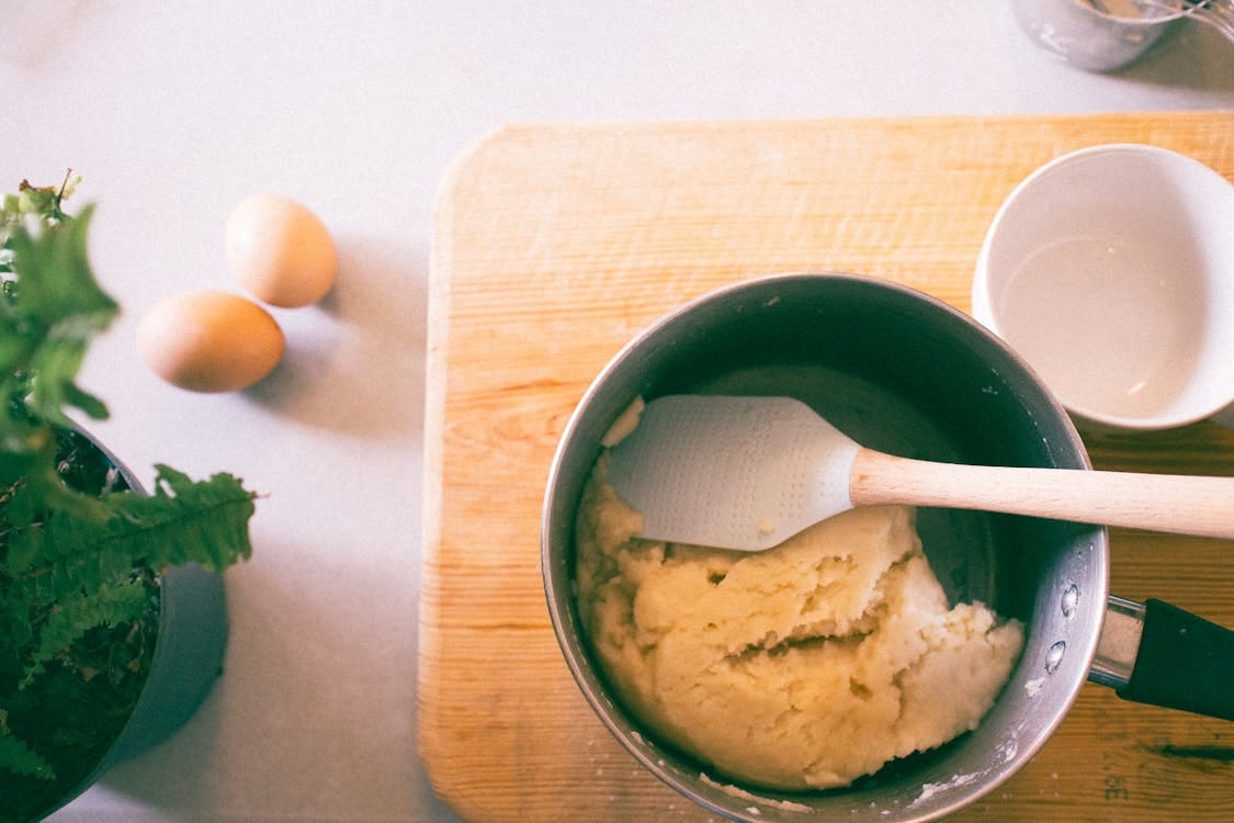 Free A kitchen setup with dough in a pot, eggs, and a spatula on a wooden board. Stock Photo Free A kitchen setup with dough in a pot, eggs, and a spatula on a wooden board. Stock Photo
