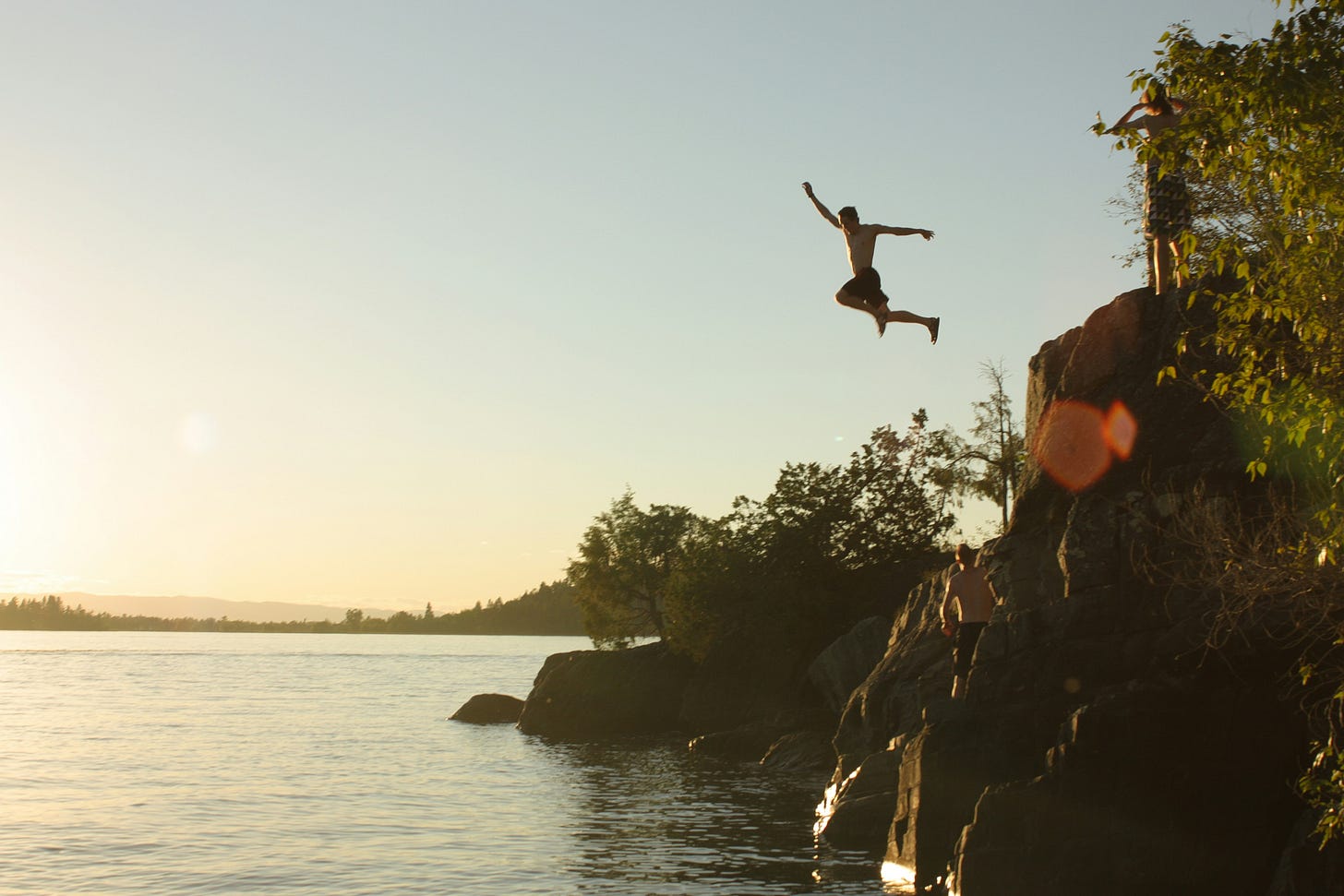 Man mid-air jumping off rocky cliff into lake at sunset, with friends watching nearby Man mid-air jumping off rocky cliff into lake at sunset, with friends watching nearby
