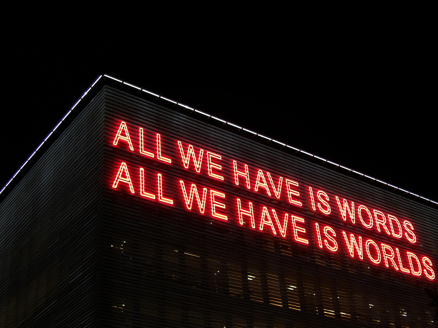 Neon sign on a building says "All we have is words. All we have is worlds."