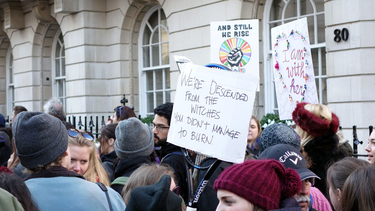 A crowd of people at a protest march, one holding a sign that says 'we're descended from the witches you didn't manage to burn'.