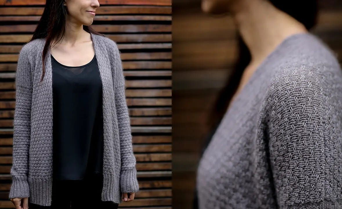 A woman stands in front of a wood wall wearing a textured gray cardigan over a black top and jeans.