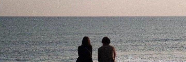 This may contain: two people standing on the beach looking out at the ocean with a kite flying in the sky