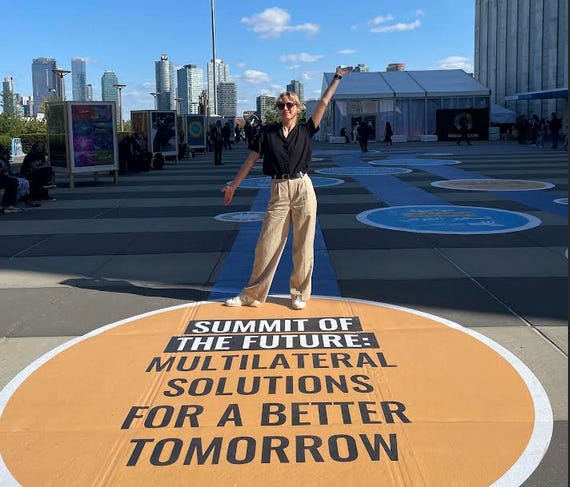 Young white blonde woman in sunglasses and a black shirt stands outside in a pavilion with a backdrop of skyscrapers, and the message written on the floor 'Summit for the Future: Multilateral solutions for a better tomorrow.'