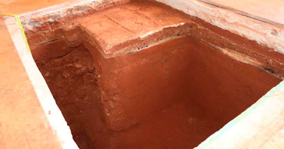 Buraco levemente retangular, com terra avermelhada. A borda é rodeada por uma faixa branca. No interior, percebe-se a demarcação de dois pisos distintos: um mais escuro e outro de tijolos, acima dele.