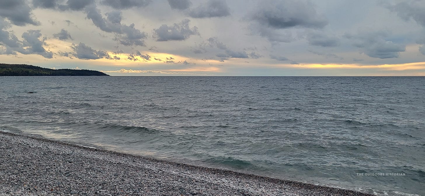 A beach covered in small, rounded stones under a choppy lake and a dramatic, sunlit sky.