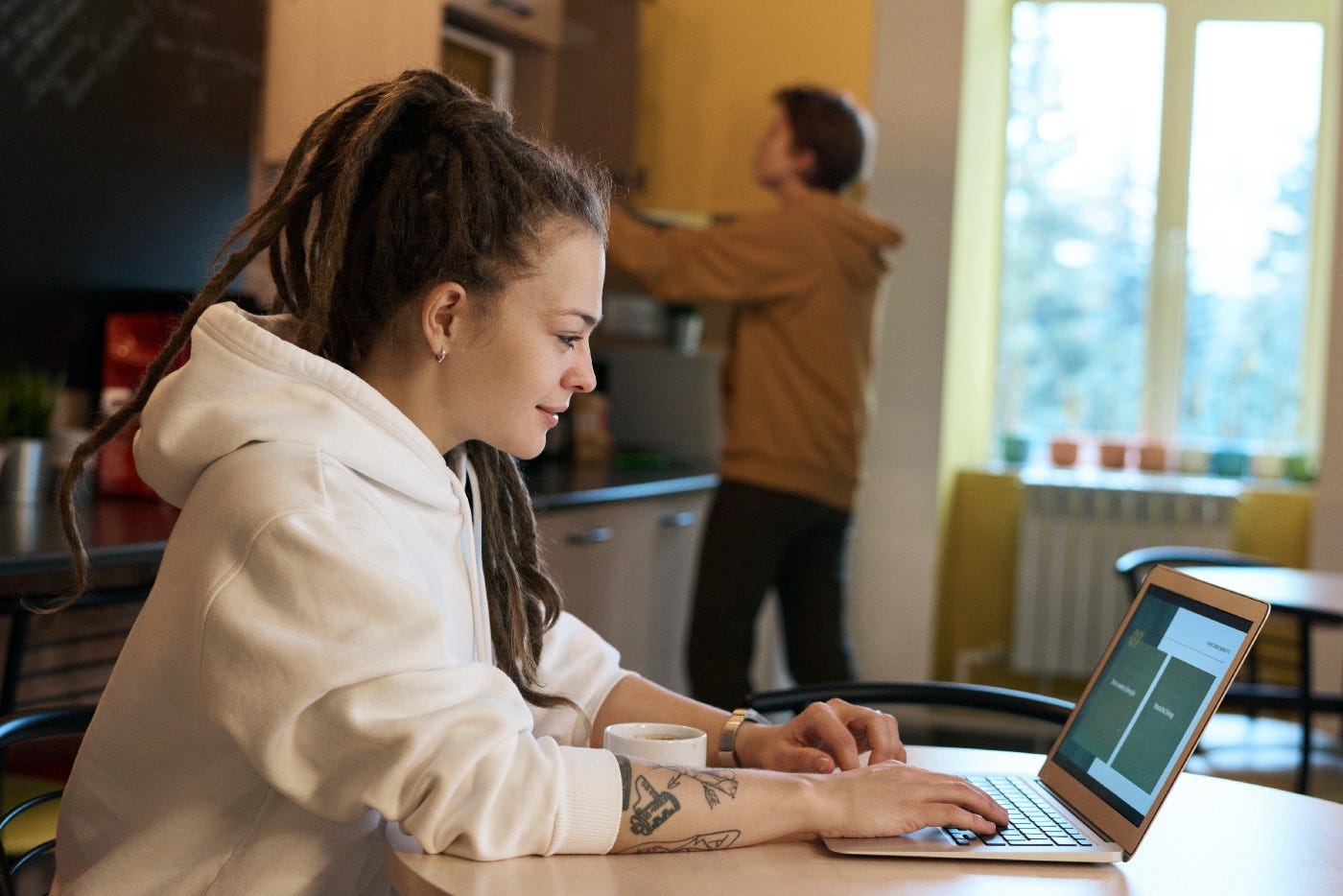 Smiling woman looking at her laptop. She sitting at a table in the kitchen while a man reaches for the microwave in the background.