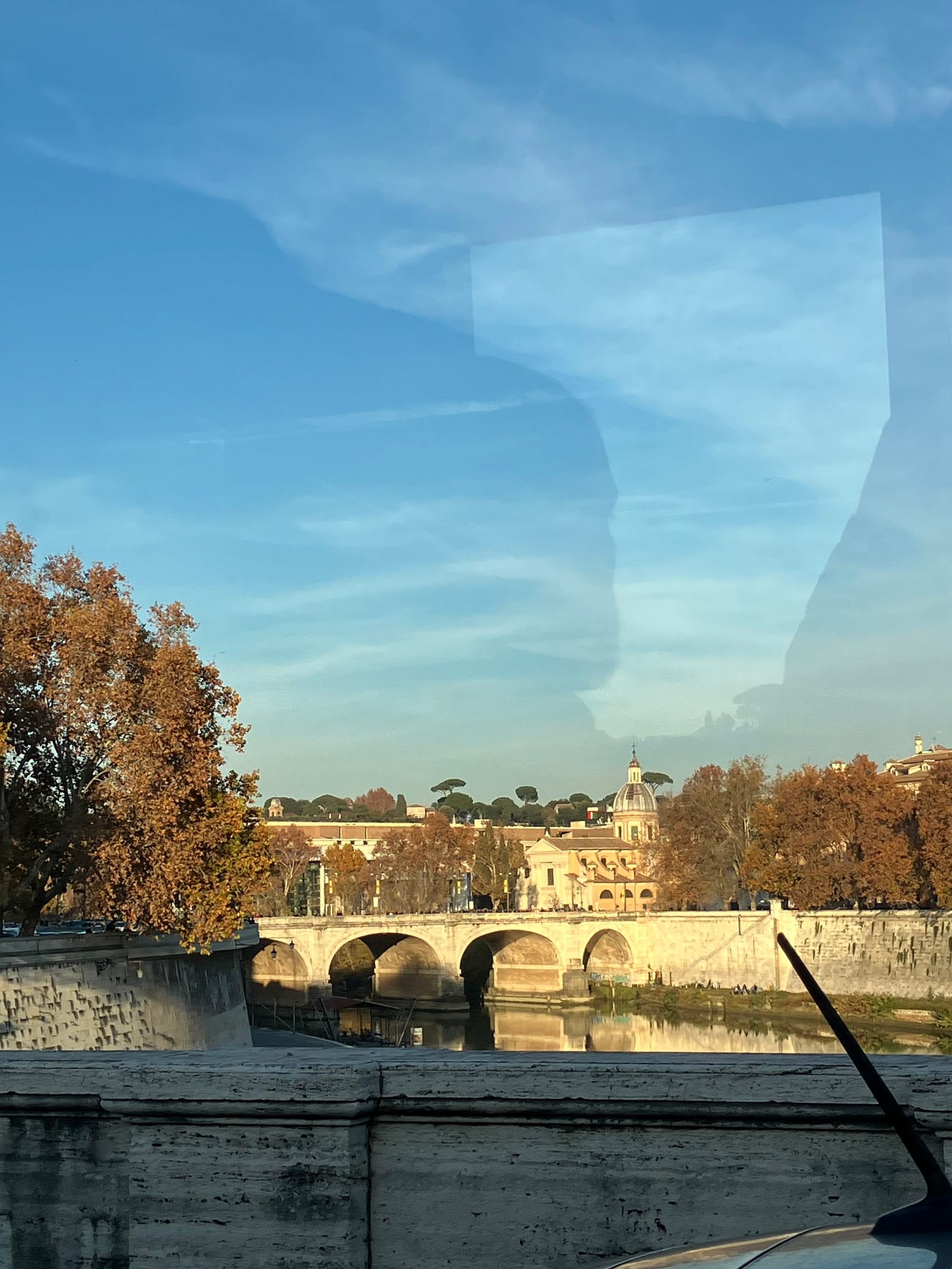 Veduta di un ponte sul fiume Tevere a Roma, con edifici storici e una cupola sullo sfondo, alberi autunnali ai lati, il cielo azzurro sopra e riflessi del paesaggio sull’acqua.
