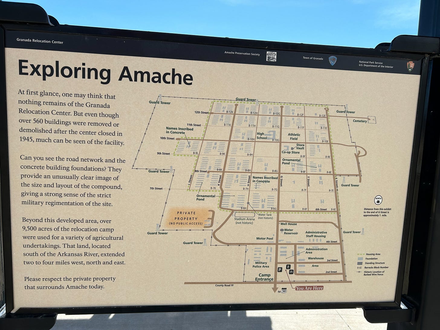 An informational plaque at the historic site titled "Exploring Amache". The plaque shows a map of the complex noting street names, building foundations and structures, boundaries, and modern visitor's facilities. Text on the lefthand column reads, "At first glance, one may think that nothing remains of the Granada Relocation Center. But even though over 560 buildings were removed or demolished after the center closed in 1945, much can be seen of the facility. / Can you see the road network and the concrete building foundations? They provide an unusually clear image of the size and layout of the compound, giving a strong sense of the strict military regimentation of the site. / Beyond this developed area, over 9,500 acres of the relocation camp were used for a variety of agricultural undertakings. That land, located south of the Arkansas River, extended two to four miles west, north and east. / Please respect the private property that surrounds Amache today".