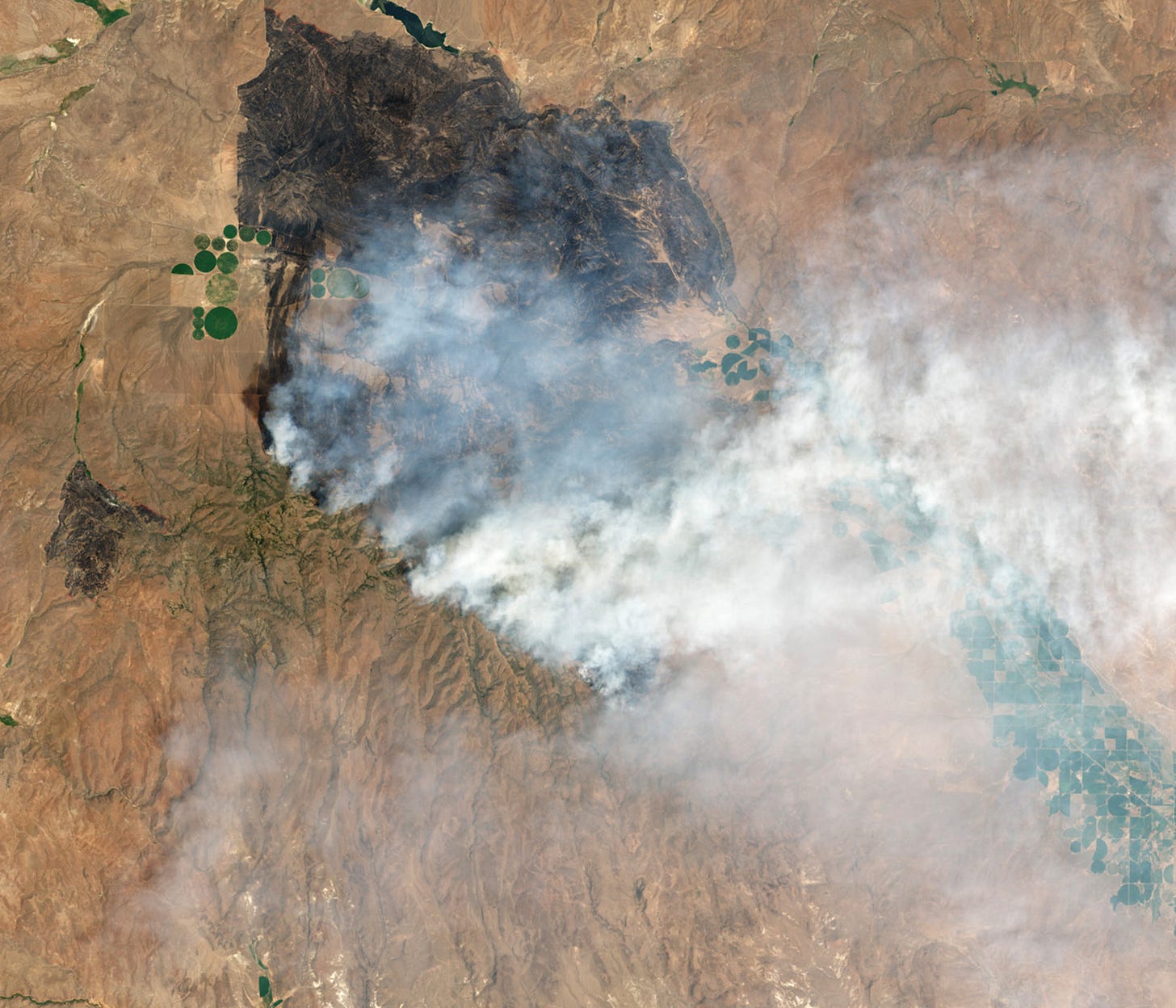 Smoke rises from one portion of a large black scar on brown earth with some green fields visible. Smoke rises from one portion of a large black scar on brown earth with some green fields visible.