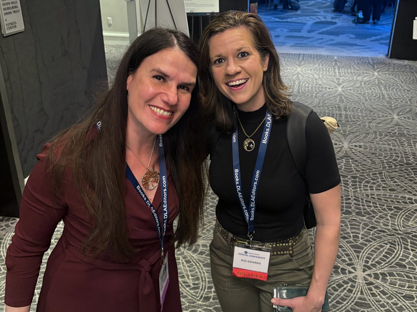 Two women (Alexandra Hidalgo and Jess Zafarris) with conference lanyards stand together looking up and smiling at the camera. They are at a conference hotel. There is a carpet with flowers on it under them. It is night time. 