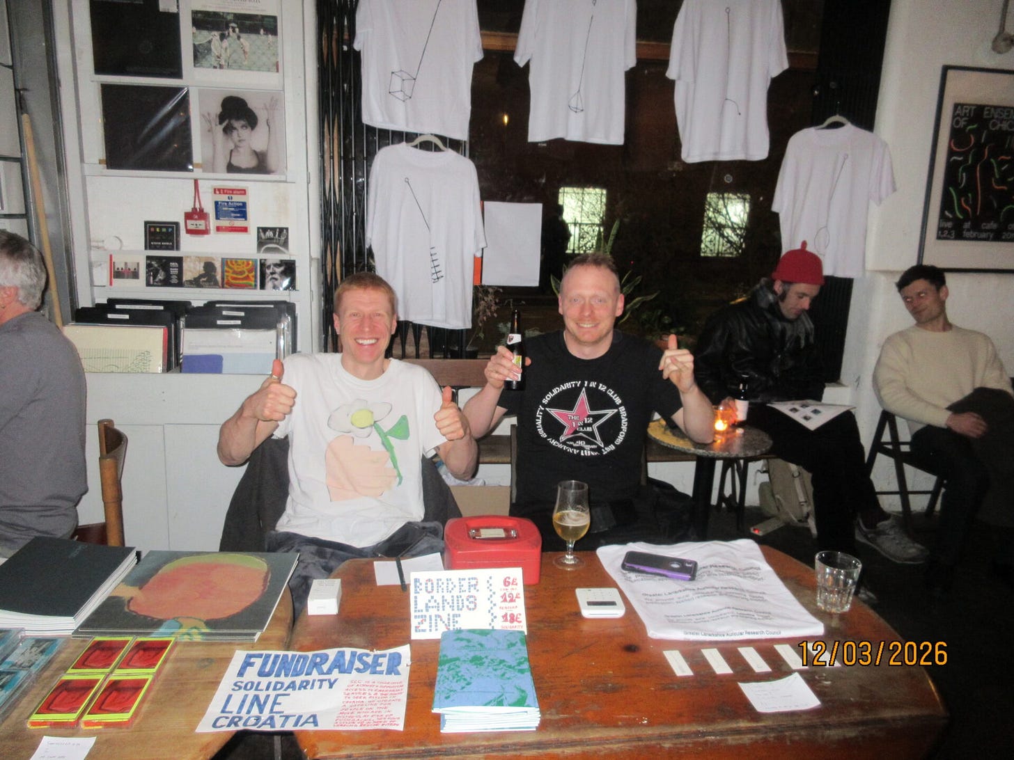 Two men in their 30s give the thumbs up while sitting at a table full of tapes, records and zines Two men in their 30s give the thumbs up while sitting at a table full of tapes, records and zines