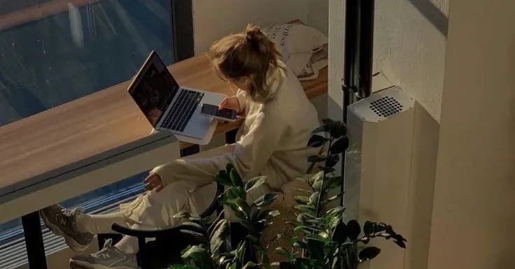 This may contain: a woman sitting at a table with a laptop computer in her lap and potted plant