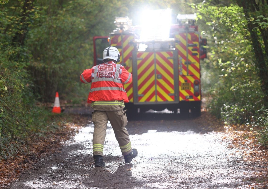 A firefighter wearing a high-visibility red jacket labeled “Tactical Advisor – Operational Assurance” stands on a muddy woodland road facing a fire engine with flashing lights and red-and-yellow chevron markings. A firefighter wearing a high-visibility red jacket labeled “Tactical Advisor – Operational Assurance” stands on a muddy woodland road facing a fire engine with flashing lights and red-and-yellow chevron markings.