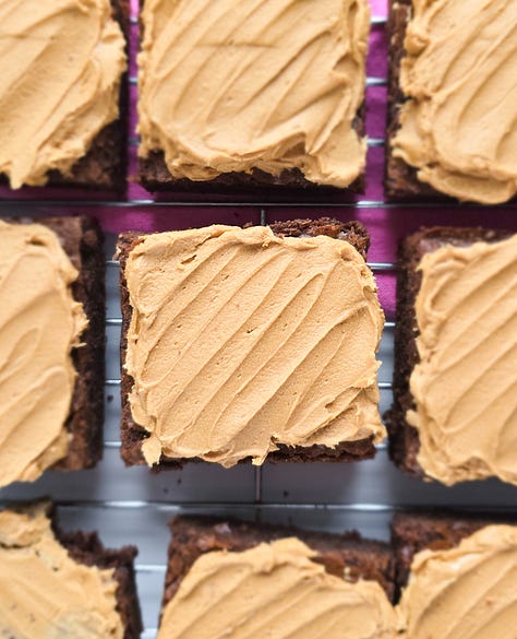 a pink baking tray with almond macaroons and glace cherries on it. A cooling rack with apple and blackberry muffins on it with apples at the top right hand of the shot. A pink cake plate with a 4 layer spiced plum upside down cake on it.