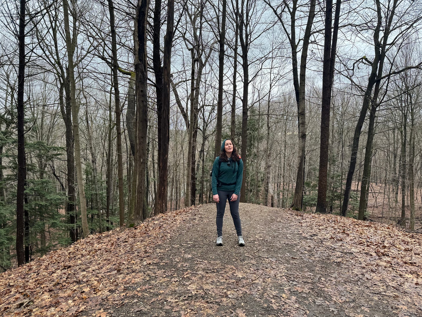 A woman in hiking gear stands on a hilltop clearing in a deciduous forest in early spring. No leaves are visible on the trees but there are plenty of dead brown ones on the ground. She looks tired. 