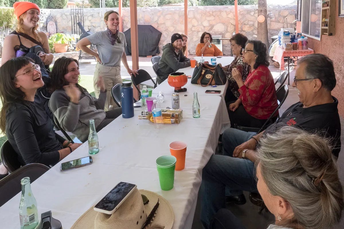 Several people sit and stand around a long table set with white tablecloth outside. They are laughing and talking while many bottles, cuts, and other objects sit on the tables, probably post-meal. Several people sit and stand around a long table set with white tablecloth outside. They are laughing and talking while many bottles, cuts, and other objects sit on the tables, probably post-meal.