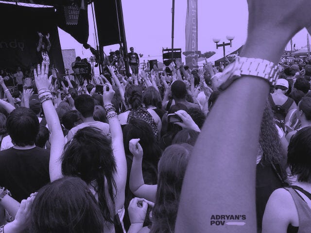 A wide-angle shot of a festival crowd at an outdoor concert. Many people in the audience have their hands raised, some making the rock sign or pointing toward the stage. The stage in the distance has a band performing, with banners and flags hanging above them. A person's arm with a white wristband is in the foreground, raised high. The image has the same desaturated, purple-tinted color overlay as the first.