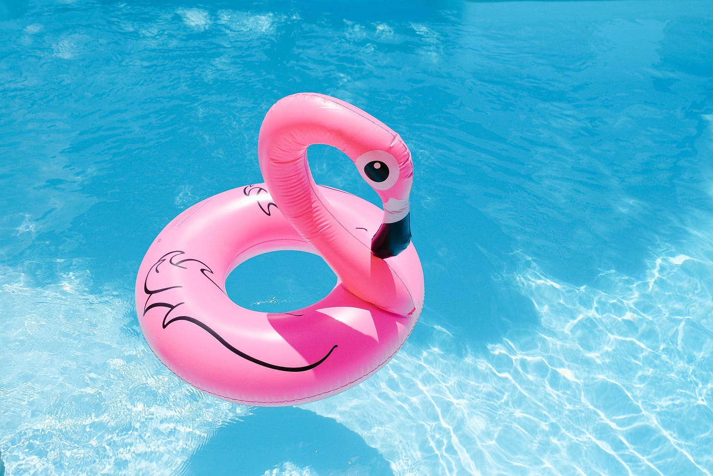 Pink flamingo pool float on bright blue water with sunlight reflections.